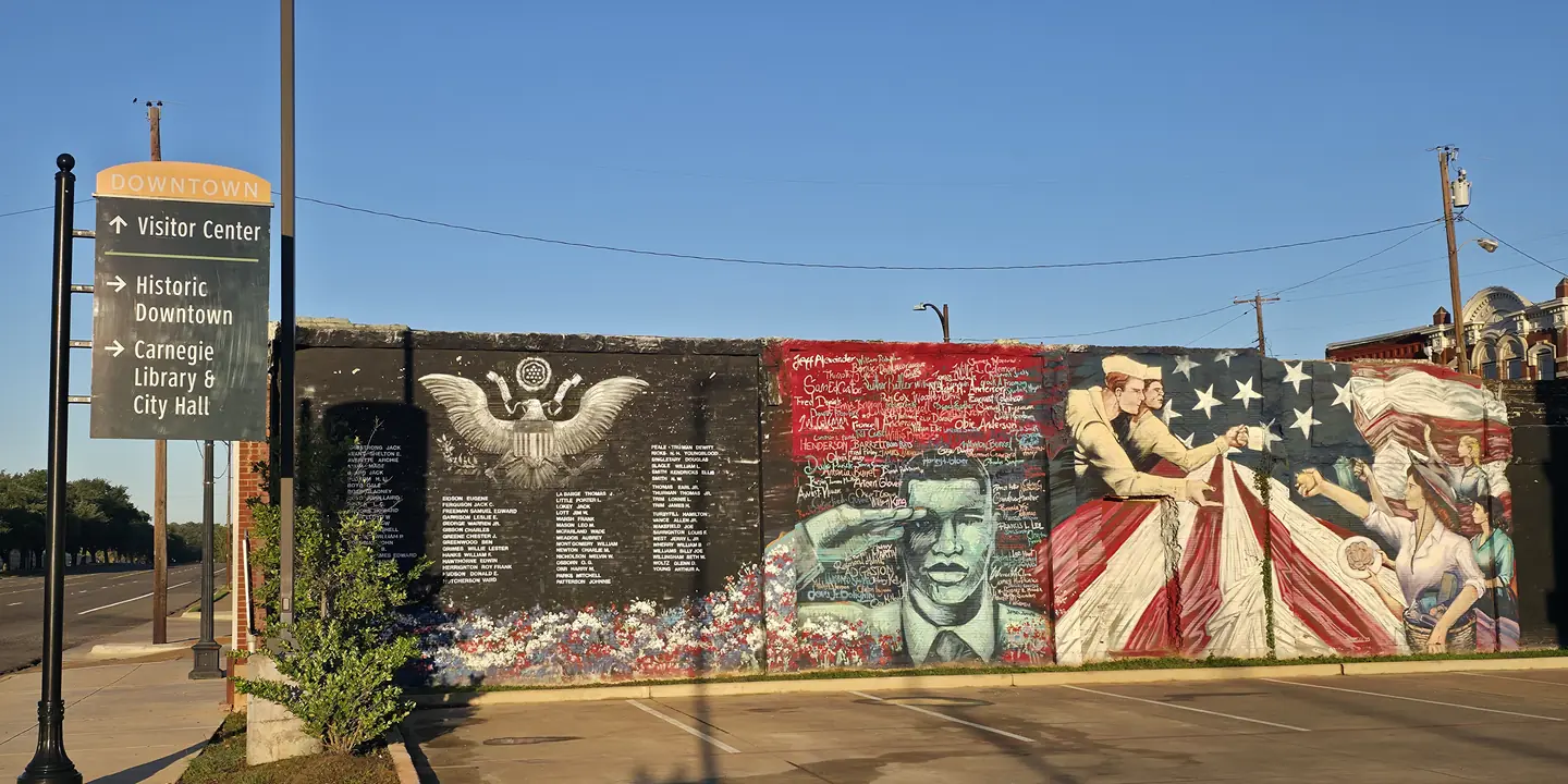 The World War II memorial mural on W. Spring Street in Palestine, Texas, with an eagle above lists of names, a saluting soldier, and sailors and women in front of an American flag. A Downtown wayfinding sign stands at the left.